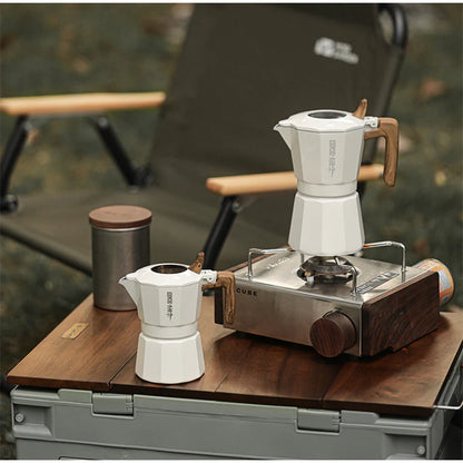 Two MHW-3BOMBER Double Valve Moka Pots with wooden handles are on a portable gas stove outdoors. A metal canister is nearby, and a green folding chair sits against a grassy backdrop, perfect for enjoying craft coffee in nature.