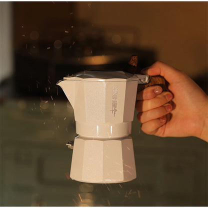 A hand holds the MHW-3BOMBER Double Valve Moka Pot 100ml, featuring a small spout and handle in white. The blurred background with sparks and warm lighting hints at its use on compatible stovetops, highlighting its durable design.