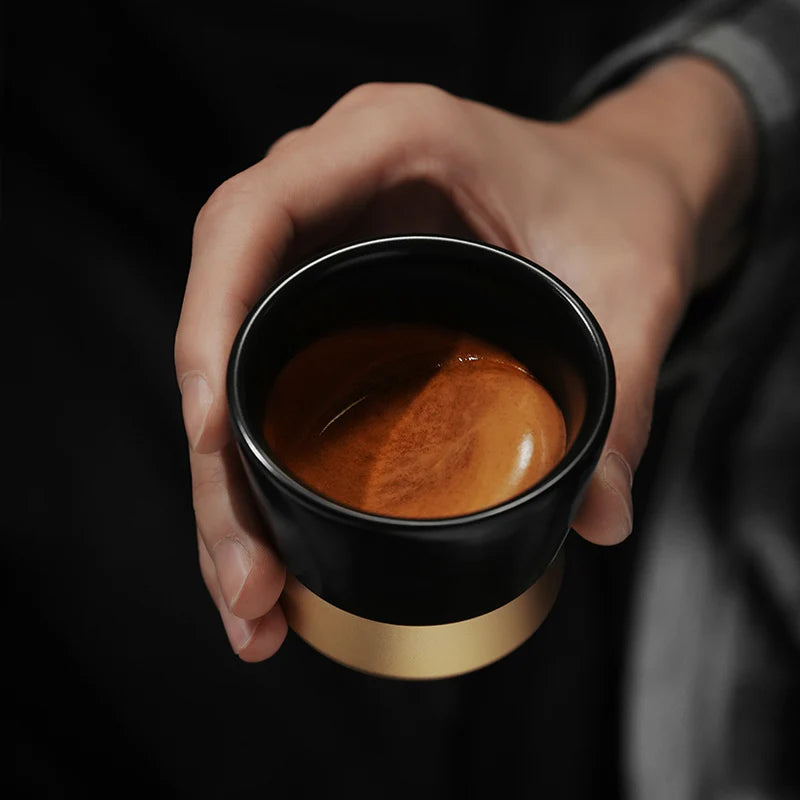 MHW-3BOMBER CERA black ceramic cup with gold base, filled with espresso. Hand holding the coffee cup.
