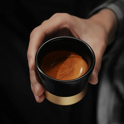 MHW-3BOMBER CERA black ceramic cup with gold base, filled with espresso. Hand holding the coffee cup.