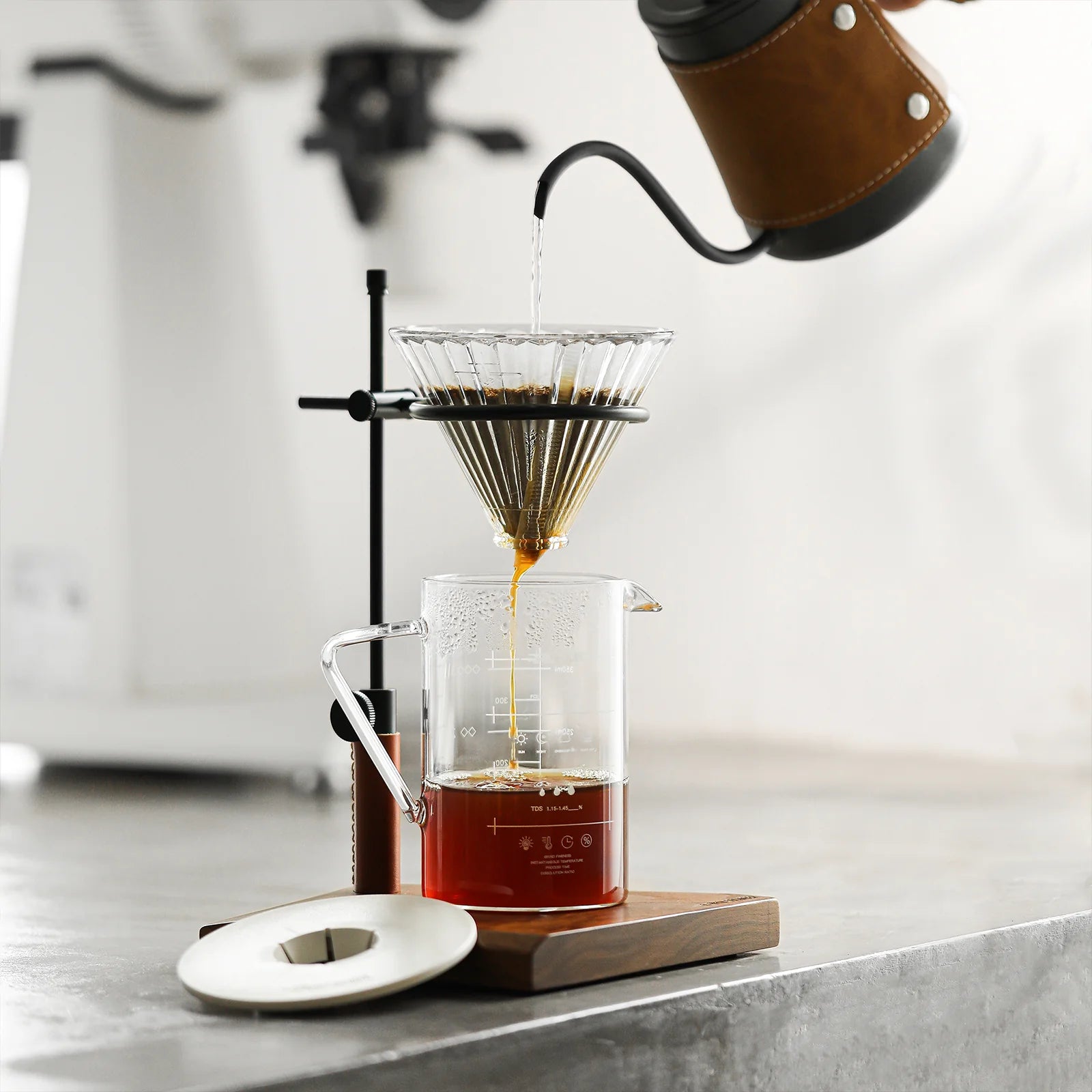 A gooseneck kettle pours hot water into the MHW-3BOMBER Elf Glass Coffee Dripper, with coffee flowing into a glass carafe on its wooden base. A reusable cone filter is used, and a white lid sits nearby.