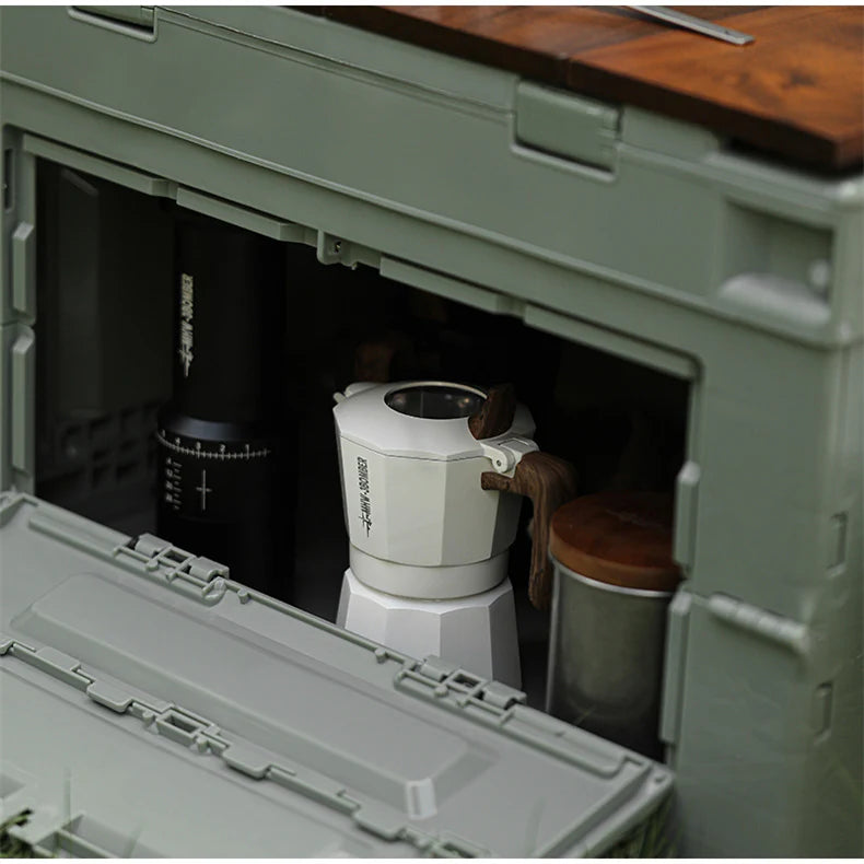 The durable green storage box with a wooden lid partially visible on top showcases an open front revealing a black cylindrical object, metal canister, and the MHW-3BOMBER Double Valve Moka Pot 100ml, ideal for craft coffee enthusiasts.