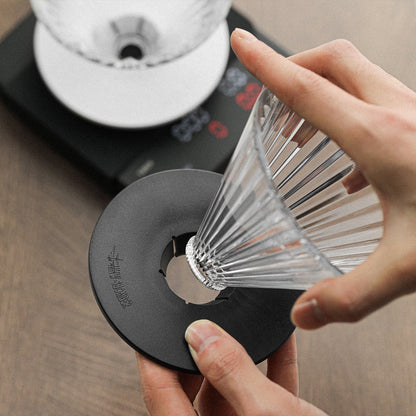 Someone assembles the MHW-3BOMBER Elf Glass Coffee Dripper on a black base. In the background, a digital scale rests on a wooden surface next to a reusable cone pour-over, ready for use.