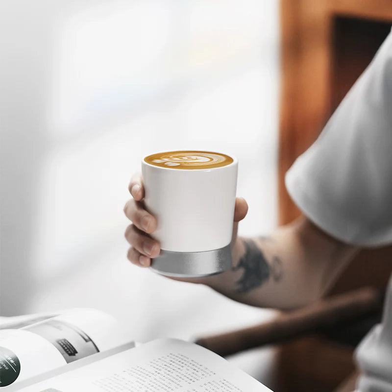 MHW-3BOMBER CERA white ceramic coffee cup with silver base and latte art. Hand holding mug over a book.