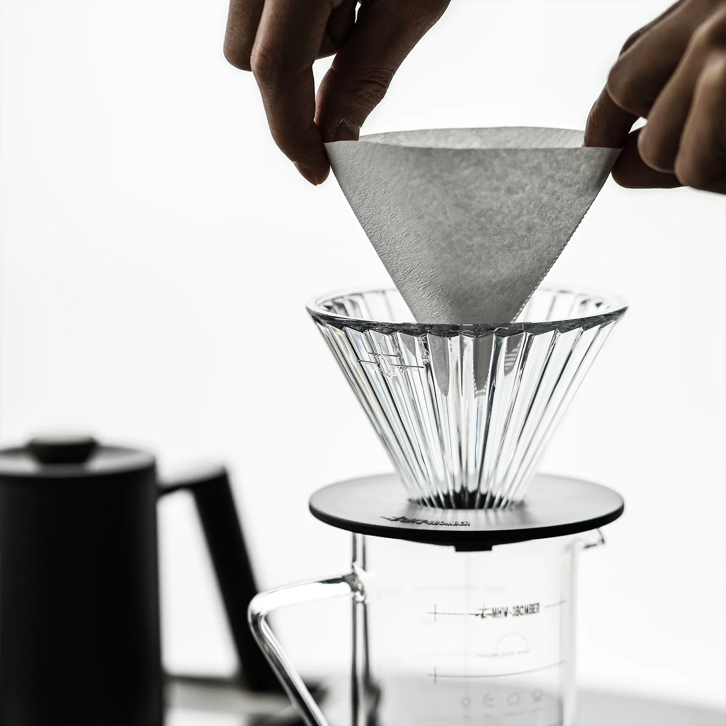 Hands gently place a conical paper filter into an MHW-3BOMBER Elf Glass Coffee Dripper above a carafe, with a black kettle poised in the background against a white backdrop.