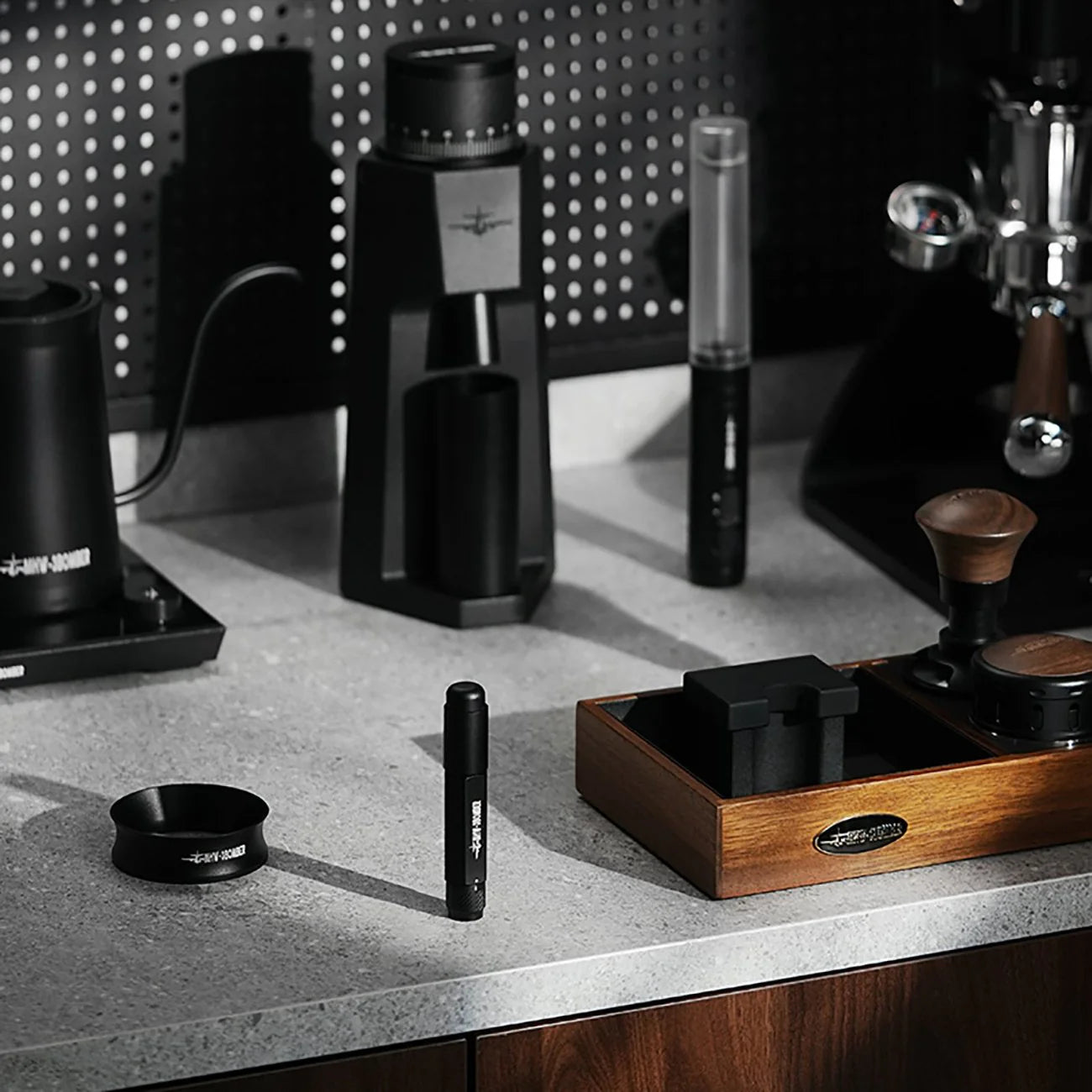 A modern kitchen countertop features coffee tools like a grinder and the MHW-3BOMBER Lightning Needle Distributor Pro WDT Tool for efficient grounds distribution. The gray surface complements the black items with wooden accents, and the well-lit setting embodies sleek organization.