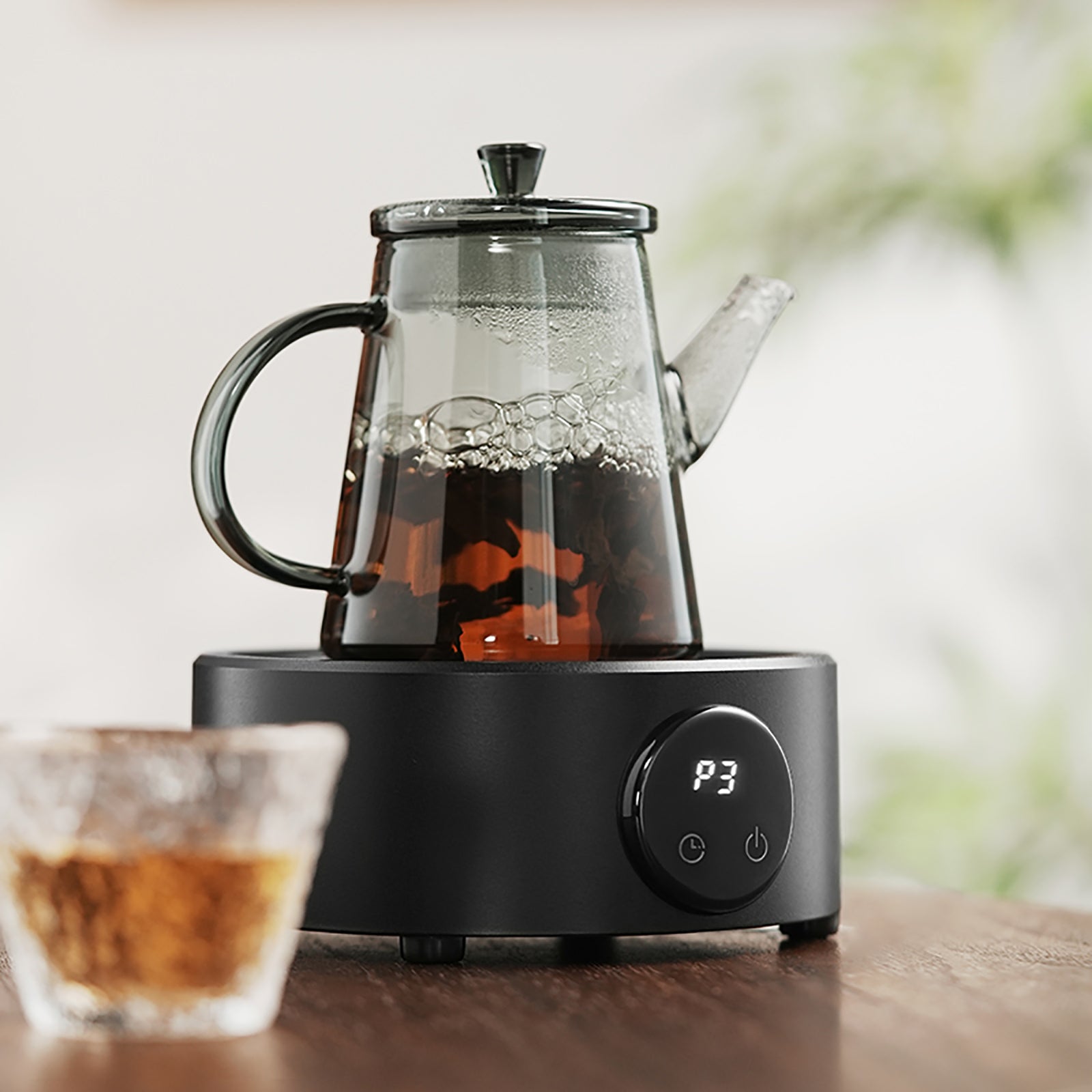 MHW-3BOMBER glass teapot with tea steeping on a black electric heater base. A clear glass cup is also visible.