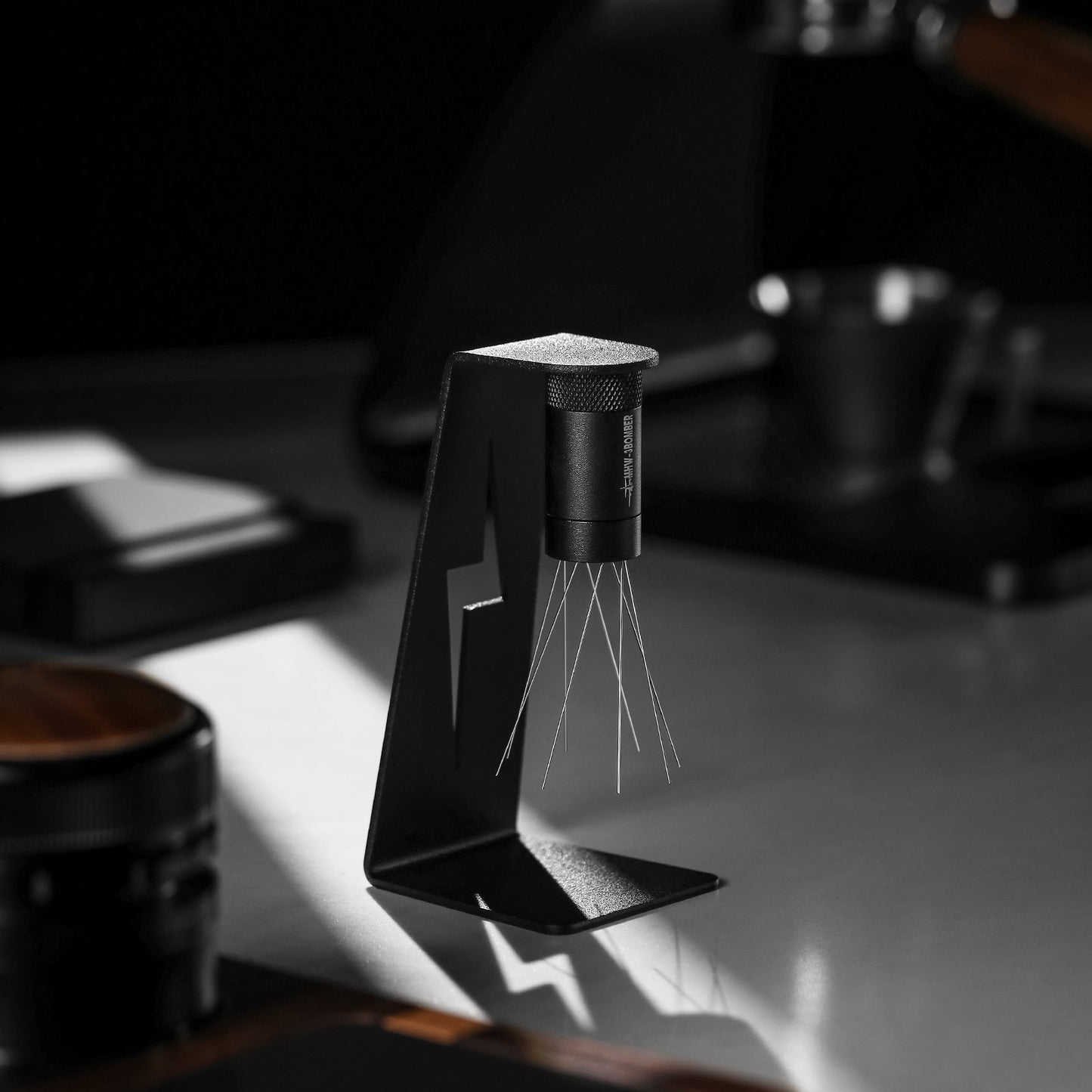 The elegant, black MHW-3BOMBER Lightning Needle Distributor WDT Tool 2.0 features multiple thin wire prongs on a sleek magnetic stand. Its styled against a blurred dark background and softly lit on a light surface to ensure optimal espresso extraction quality.