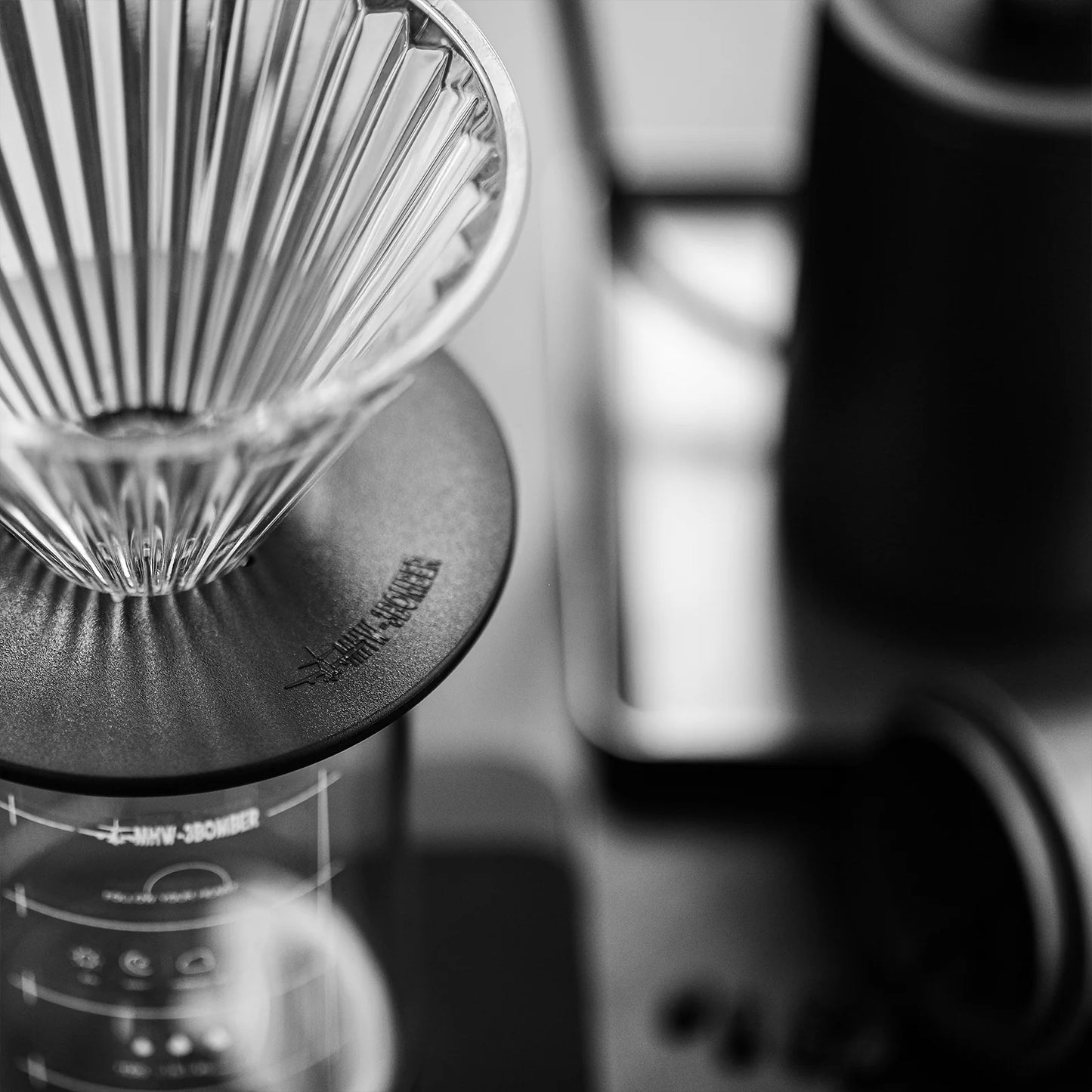 A black and white close-up features an MHW-3BOMBER Elf Glass Coffee Dripper with a ridged design on a carafe, alongside a reusable cone filter. A scale is partially visible underneath, while blurred background elements include a kettle and tray.