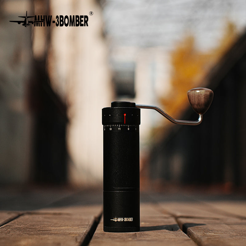 The MHW-3BOMBER Blade R3 Manual Coffee Grinder features a sleek black cylindrical design with a stainless steel burr, metal crank handle with a wooden knob, and an airplane logo. It stands on wood against a blurred outdoor backdrop.