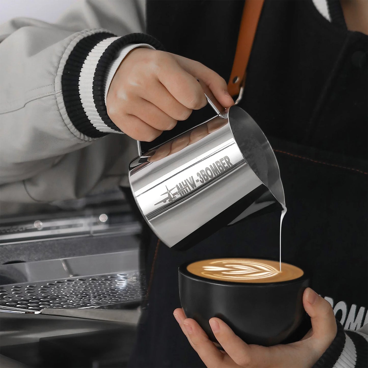 Wearing a black and gray jacket and a dark apron, a person uses the [Pre-Order] MHW-3BOMBER Latte Art Pitcher 5.0 Glossy to pour steamed milk into a black cup with latte art, standing by the coffee machine.