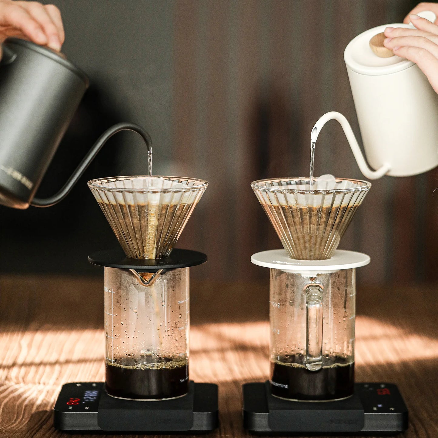 Two people pour hot water from kettles into reusable cone pour-overs with glass carafes on separate digital scales. The left kettle is black, the right is white, and an MHW-3BOMBER Elf Glass Coffee Dripper with a triangular diversion groove ensures perfect coffee brewing into the carafes below.