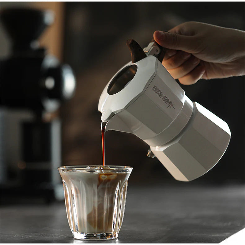 A hand pours coffee from an MHW-3BOMBER Double Valve Moka Pot 100ml into a glass with milk, creating a marbled effect. A blurred coffee grinder is in the background, highlighted by its compatibility with all stovetops and durable design.