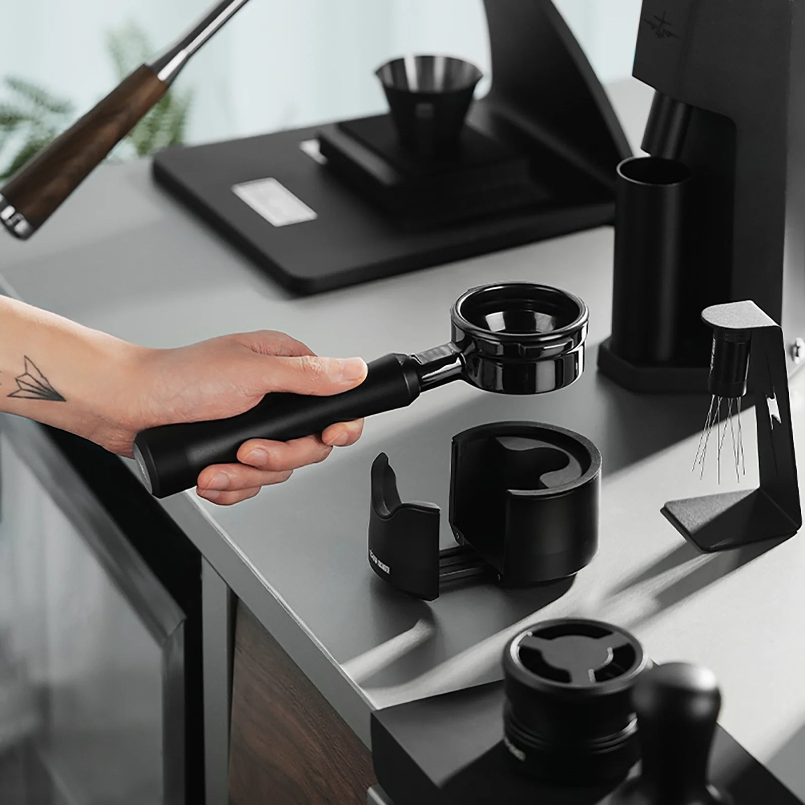 MHW-3BOMBER black bottomless portafilter held on counter with espresso machine accessories.