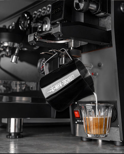 A close-up of a coffee machine pouring milk from the [Pre-Order] MHW-3BOMBER Latte Art Pitcher 5.0 Glossy into espresso, crafting a layered drink, with the machines dials and black exterior visible in the background.