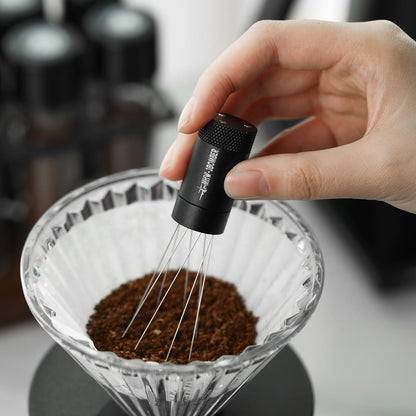 A hand expertly uses the MHW-3BOMBER Lightning Needle Distributor WDT Tool 2.0, featuring thin wires, to stir ground coffee in a glass dripper and enhance extraction quality. In the blurred background, coffee equipment rests on a sleek magnetic stand.