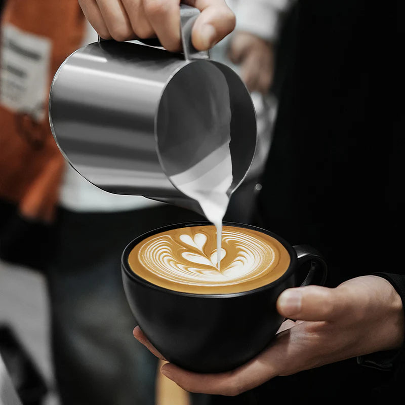 MHW-3BOMBER milk pitcher pouring latte art. Stainless steel pitcher, black coffee cup, heart design in foam.