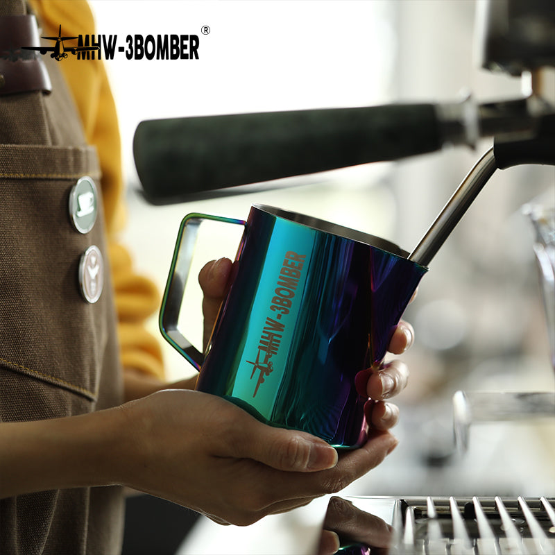 The apron-clad person expertly holds a colorful metallic MHW-3BOMBER Latte Art Pitcher 5.0 near an espresso machine steam wand, using the crocodile spout for precision in crafting a perfect cup of coffee.