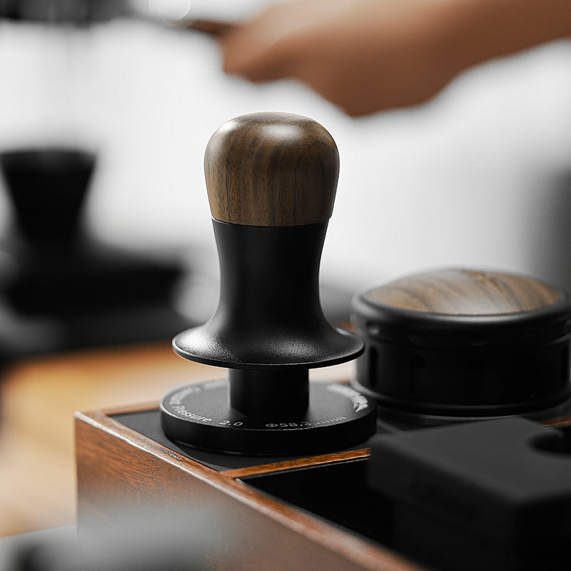 A close-up of the MHW-3BOMBER Flash Tamper featuring an ergonomic wooden handle and black base on a wooden surface. The blurred background shows a hand with a tool. Its three-spring design is next to a sleek black box with a wooden top, providing 30lbs constant pressure and sound feedback.