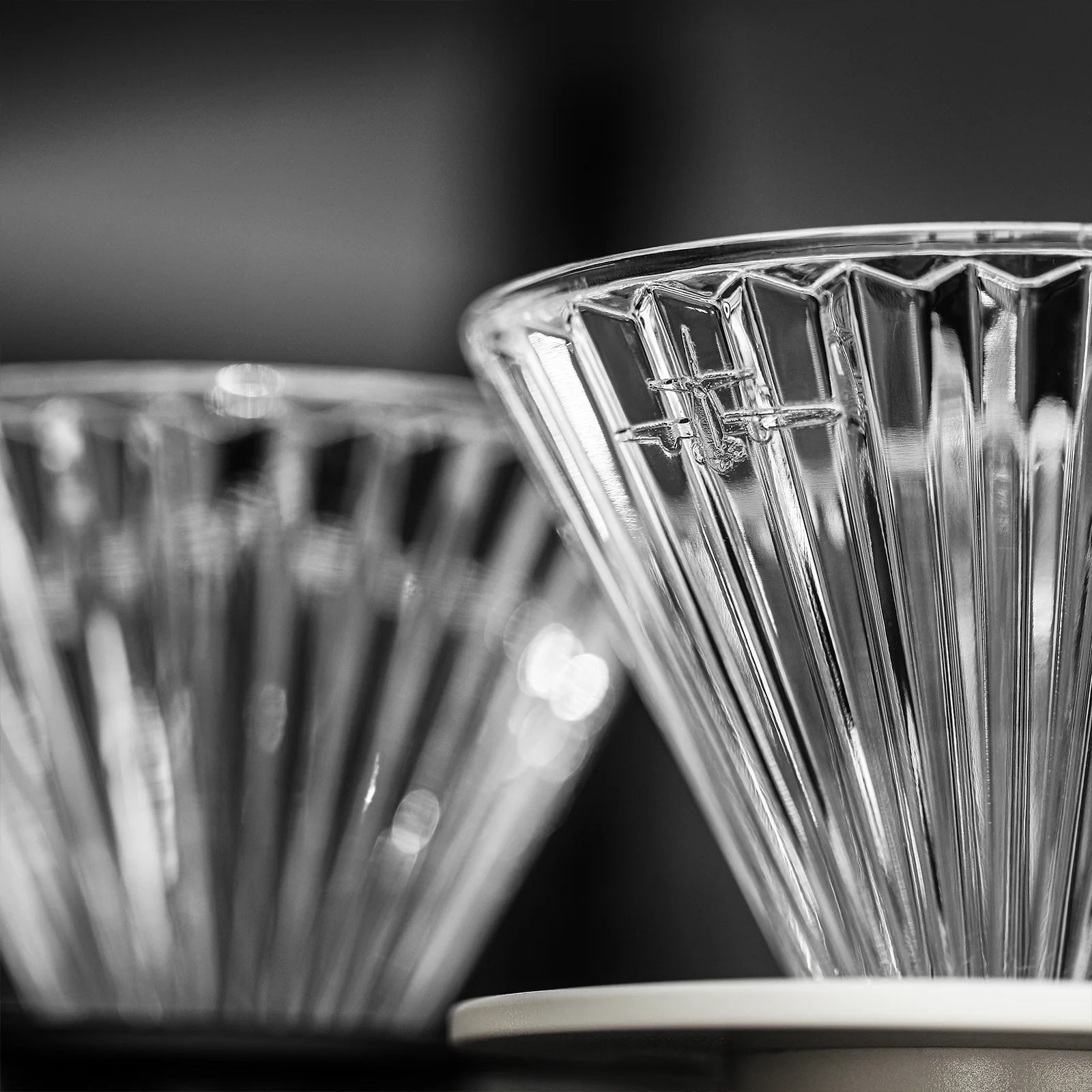 Close-up of a MHW-3BOMBER Elf Glass Coffee Dripper with triangular grooves against a blurred backdrop. Another dripper is visible, enhancing the symmetrical and reflective design, ideal for sustainable brewing with a reusable cone pour-over.