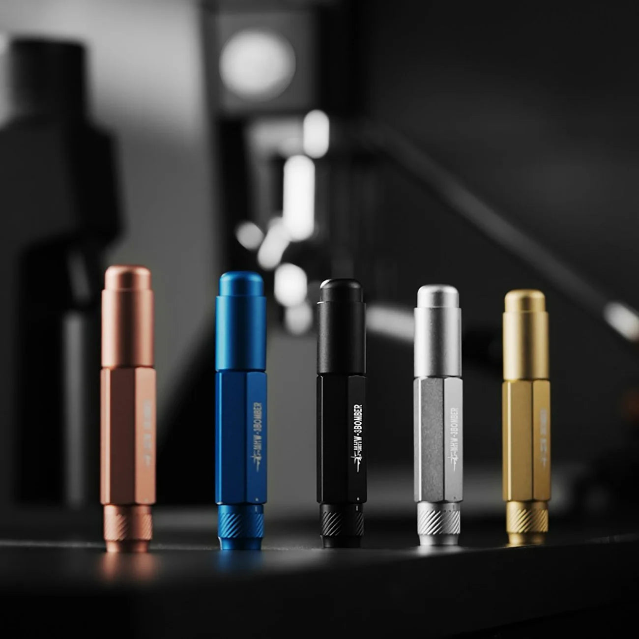 Five MHW-3BOMBER Lightning Needle Distributor Pro WDT Tools feature a metallic telescopic design in copper, blue, black, silver, and gold. Theyre neatly lined on a dark surface with a blurred background suggesting a modern, minimalistic setting.