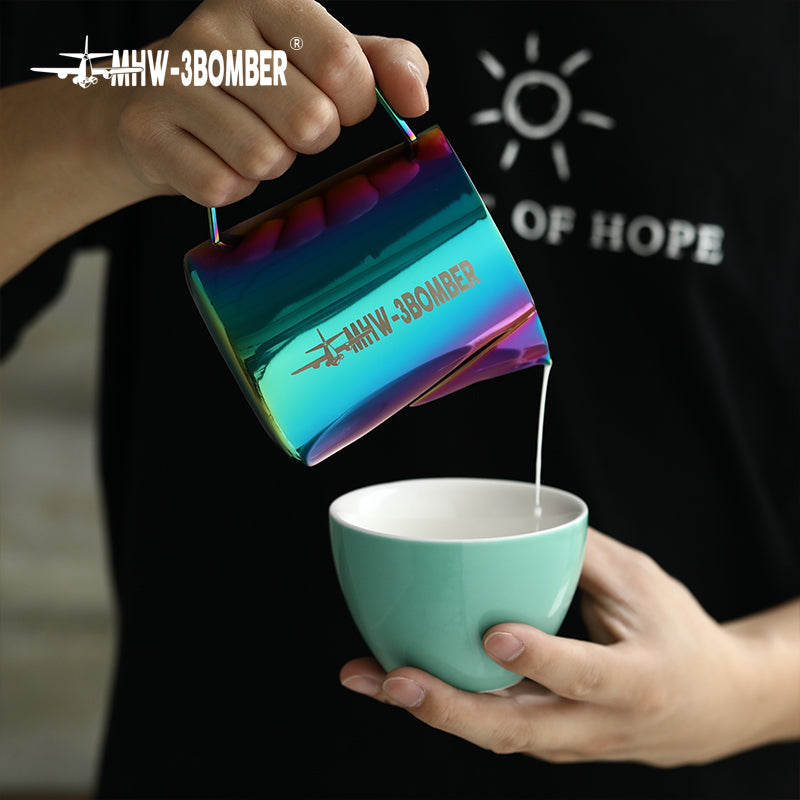 A person is pouring liquid from an MHW-3BOMBER Latte Art Pitcher 5.0 Colorful, featuring a crocodile spout and an airplane logo, into a light blue mug. A sun and OF HOPE are visible in the background on a black surface.