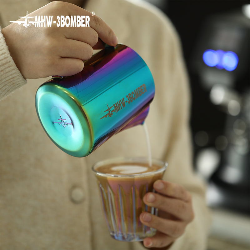 A person pours milk from the MHW-3BOMBER Latte Art Pitcher 5.0 Colorful into a clear glass of coffee, its sleek crocodile spout and MHW-3BOMBER lettering making it stand out against the blurred backdrop of a coffee machine.