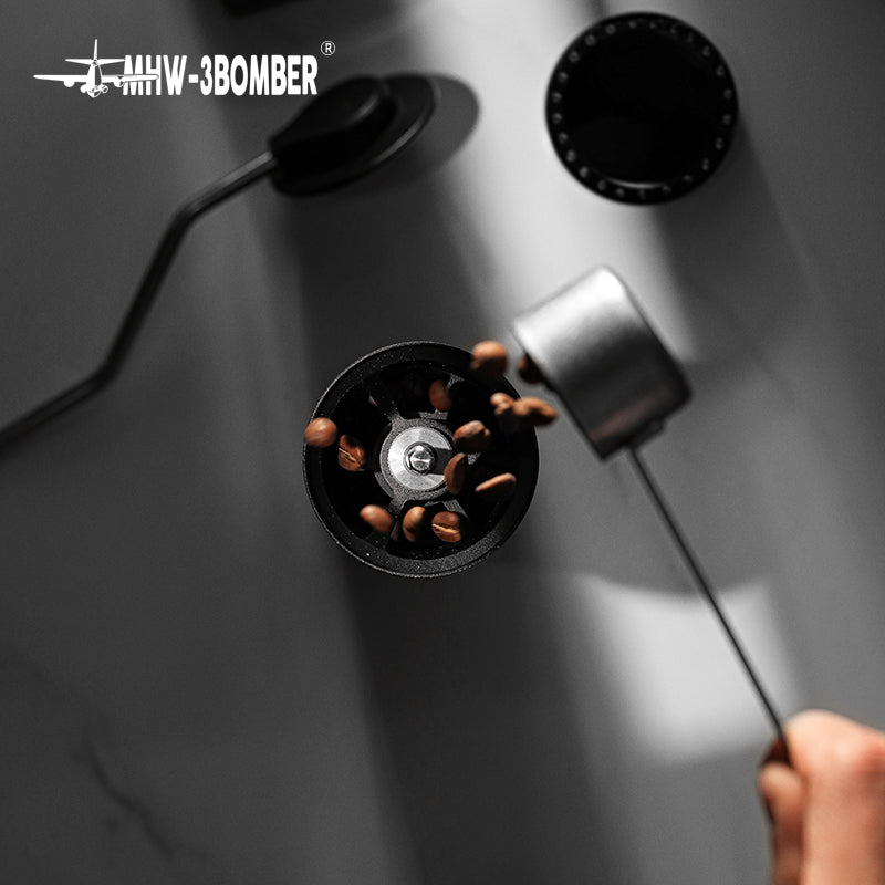 An overhead shot captures MHW-3BOMBERs Blade R3 Manual Coffee Grinder on a white surface as coffee beans are poured from a metallic scoop. The dramatic shadows, small black circular lids, and stainless steel burr enhance the visual appeal.