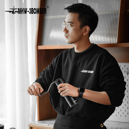 A person wearing glasses and a black sweatshirt with MHW-3BOMBER grinds coffee beans using the MHW-3BOMBER Blade R3 Manual Coffee Grinder with stainless steel burrs. Theyre indoors near a window, with a shelf of jars in the background.