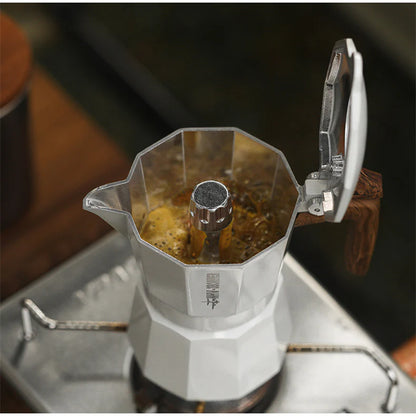 The MHW-3BOMBER Double Valve Moka Pot 100ml, with a durable wooden handle and knob, sits on a stove brewing coffee. This silver craft coffee maker rests on a simple gas burner, compatible with various stovetops for convenience.