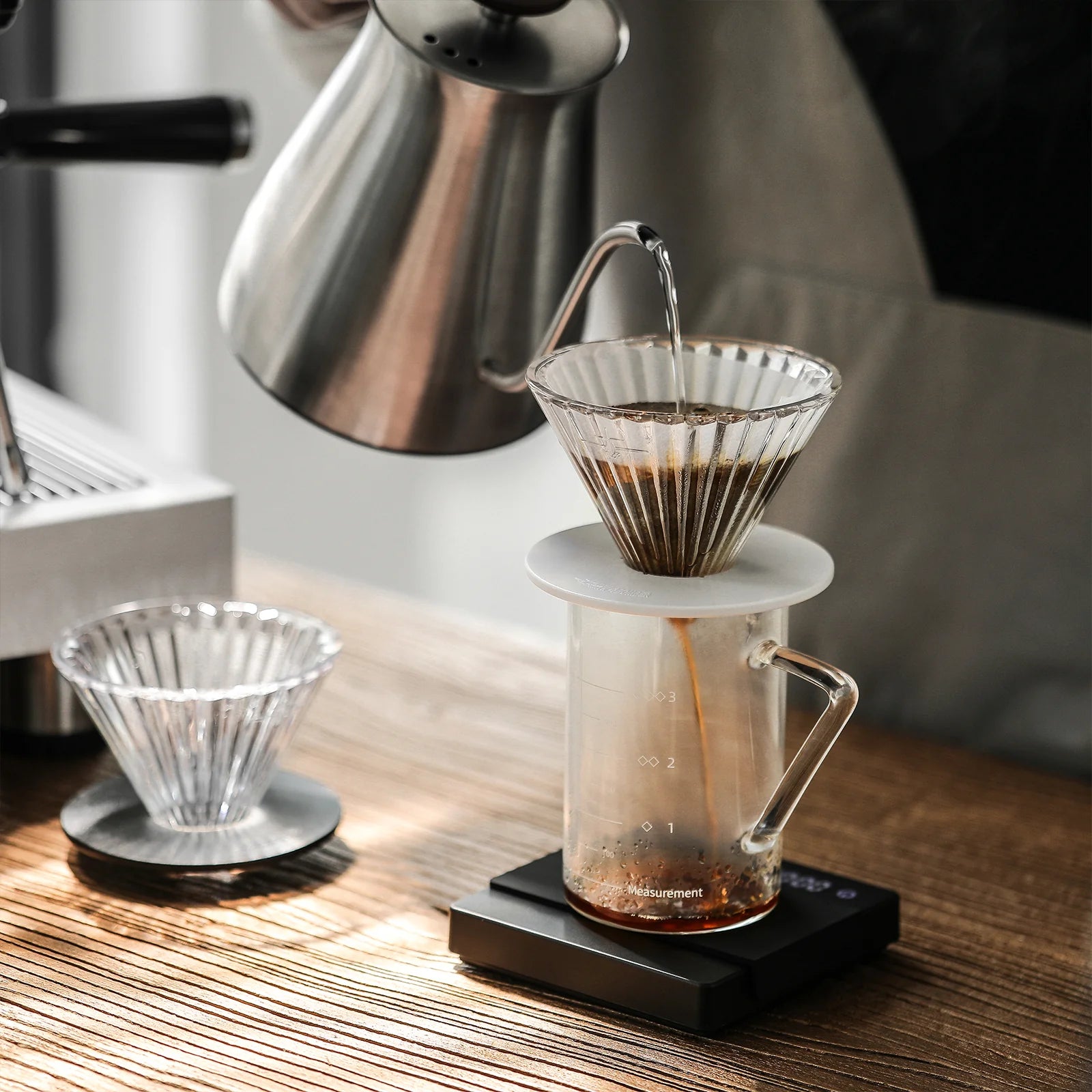 Pouring hot water from a silver kettle into the MHW-3BOMBER Elf Glass Coffee Dripper on a glass cup, this elegant setup on a wooden table with a digital scale underneath, and another dripper with a reusable cone filter nearby.