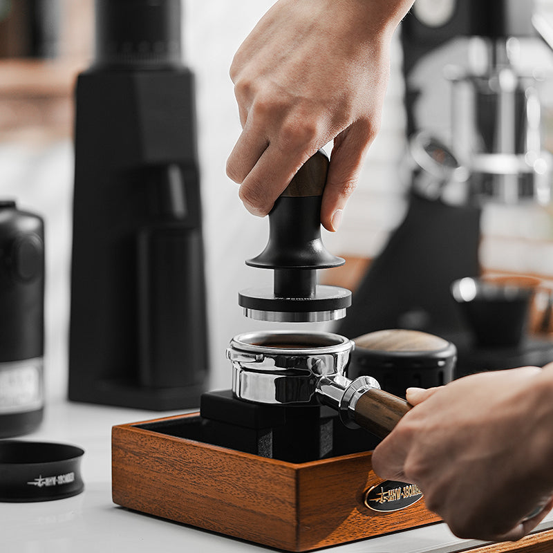 Someone is using the MHW-3BOMBER Flash Tamper with a 30lbs constant pressure and ergonomic handle to tamp coffee grounds in a portafilter held over a wooden box, with blurred coffee equipment in the background.