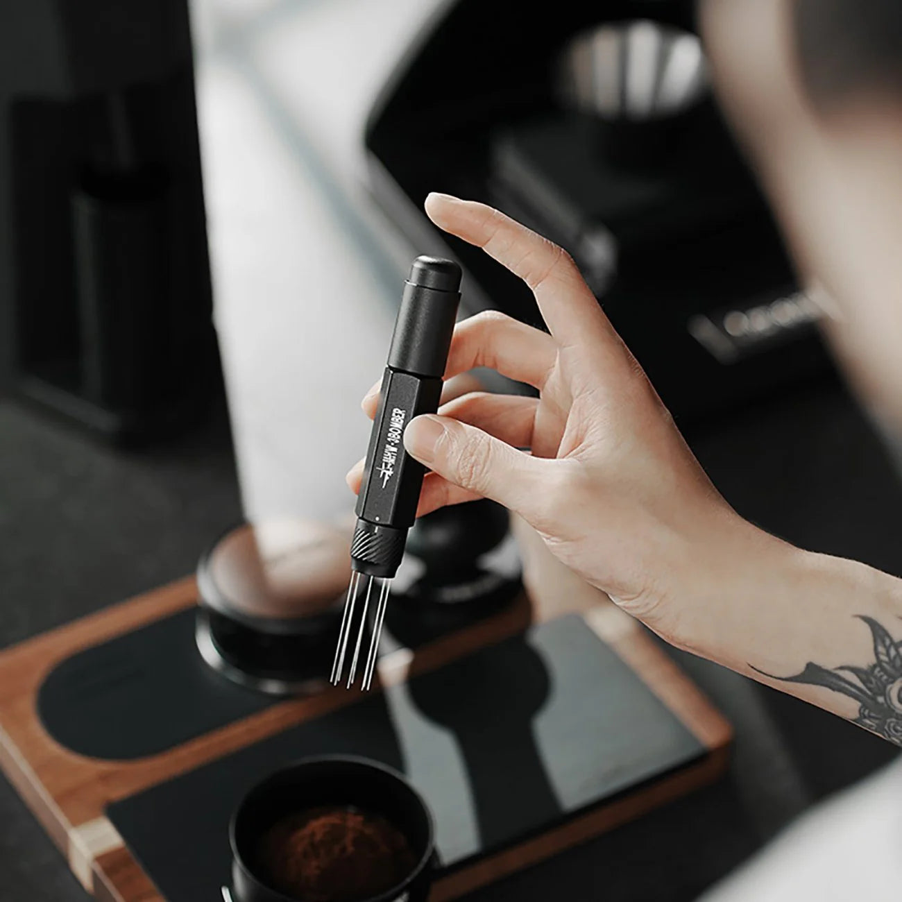 A person is seen holding a black MHW-3BOMBER Lightning Needle Distributor Pro WDT Tool with metal prongs and a matte finish, standing over a coffee station complemented by wooden and black components, part of an efficient setup for perfect brew distribution.