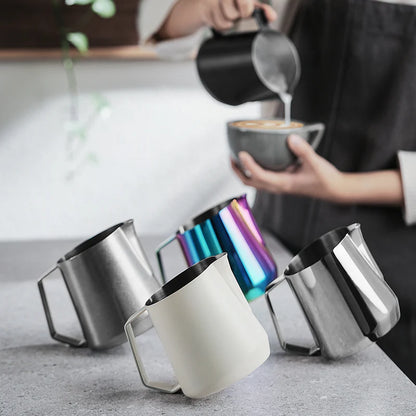 MHW-3BOMBER GT milk pitchers in various colors (stainless, white, rainbow) on a counter. Milk being poured from black pitcher.