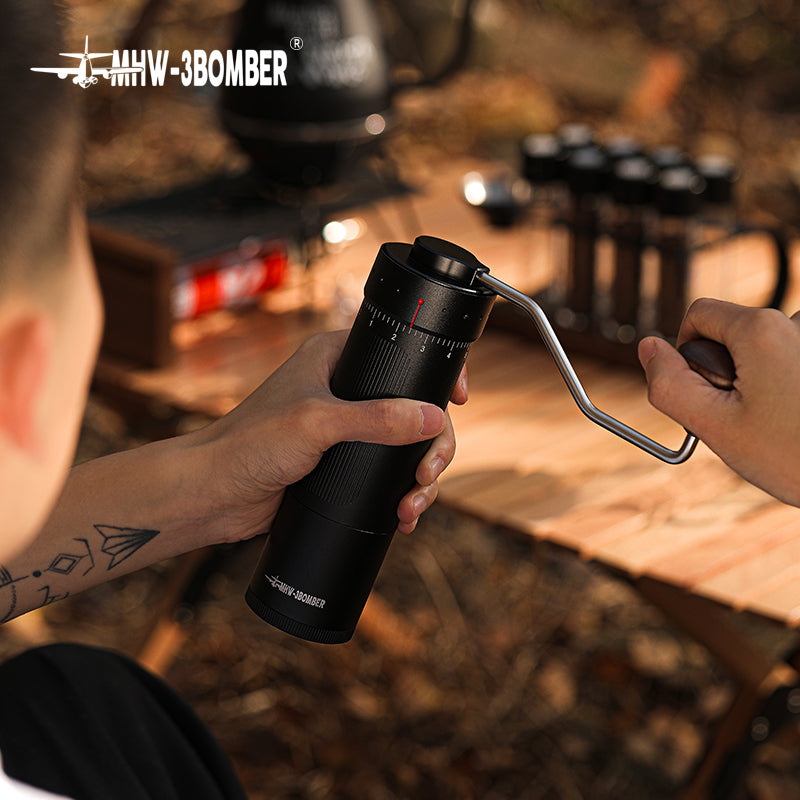 A person uses the MHW-3BOMBER Blade R3 Manual Coffee Grinder on a wooden table outdoors, with blurred camping gear in the background. Their small tattoo adds character to the serene setting as they enjoy a precise coffee-making experience with its stainless steel burr.