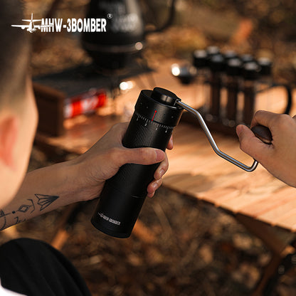 A person uses the MHW-3BOMBER Blade R3 Manual Coffee Grinder on a wooden table outdoors, with blurred camping gear in the background. Their small tattoo adds character to the serene setting as they enjoy a precise coffee-making experience with its stainless steel burr.