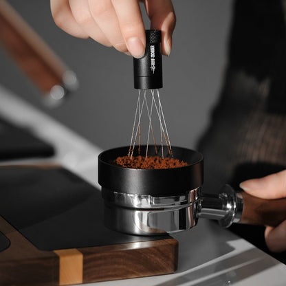 A close-up shows a hand using the MHW-3BOMBER Lightning Needle Distributor WDT Tool 2.0 with metal prongs to evenly spread coffee grounds in a portafilter, enhancing extraction quality. The tool has a black handle, while the wooden-handled portafilter rests on a modern countertop next to its magnetic stand.