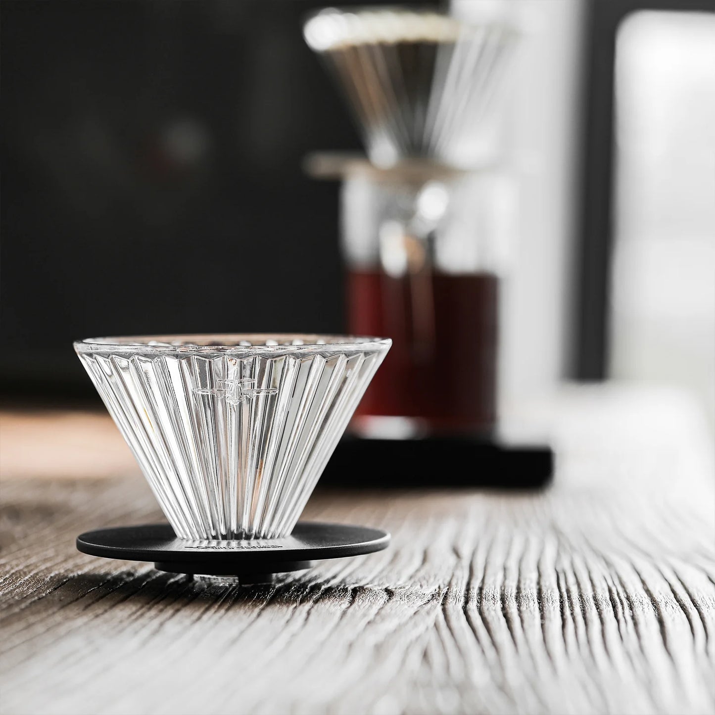 A MHW-3BOMBER Elf Glass Coffee Dripper rests on a wooden table while a reusable cone pour-over drips into a coffee-filled cup. Natural light from a nearby window beautifully illuminates the scene.