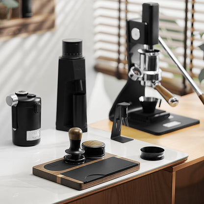 A modern coffee setup on a counter features a manual espresso machine, the MHW-3BOMBER Flash Tamper with 30lbs constant pressure and sound feedback, a coffee grinder, and a tamping mat. The minimalist workspace is bathed in natural light filtering through blinds in the background.