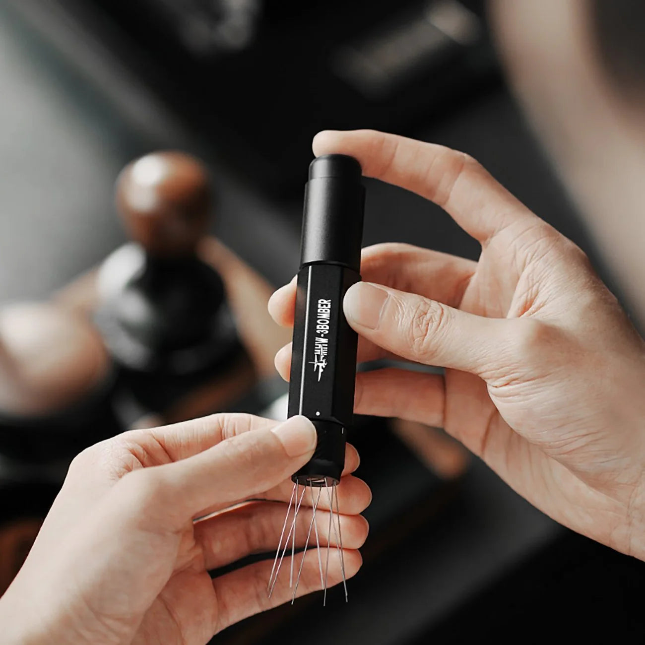 A person holds the sleek black MHW-3BOMBER Lightning Needle Distributor Pro WDT Tool, featuring an innovative telescopic design. It bears white text, with a blurred kitchen background adding charm to this essential tool.