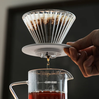 A hand delicately holds the MHW-3BOMBER Elf Glass Coffee Dripper on a carafe, expertly brewing coffee. The transparent, reusable cone pour-over design ensures a smooth flow of rich brown liquid into the vessel below, creating the perfect morning ritual.