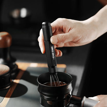 A person uses the MHW-3BOMBER Lightning Needle Distributor Pro WDT Tool to stir a small dark bowl, showcasing efficient grounds distribution. Only their hand is visible against a dimly lit kitchen with blurred objects in the background.