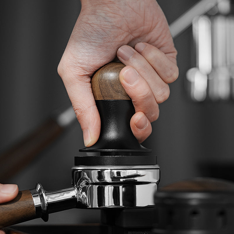 A hand uses the MHW-3BOMBER Flash Tamper, which features ergonomic design and 30lbs constant pressure, to press espresso grounds in a metallic portafilter. The focus is on the tamper and portafilter against a softly blurred background.