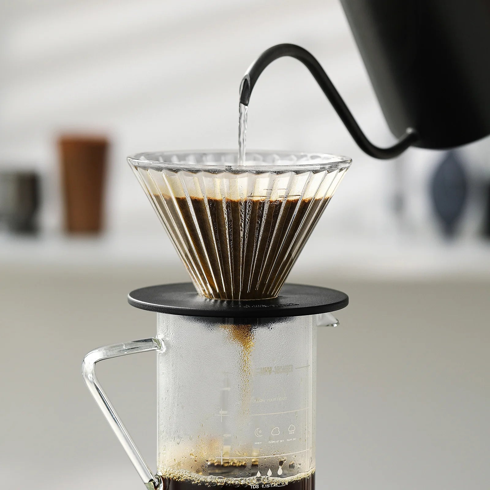 The MHW-3BOMBER Elf Glass Coffee Dripper with a reusable cone pour-over sits on a glass carafe as hot water pours from a black gooseneck kettle. Coffee flows through the dripper’s triangular diversion groove against a softly blurred background, creating a serene scene.