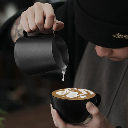 MHW-3BOMBER GT Milk Pitcher in use. Black pitcher pouring milk into black cup with latte art. Barista visible. 