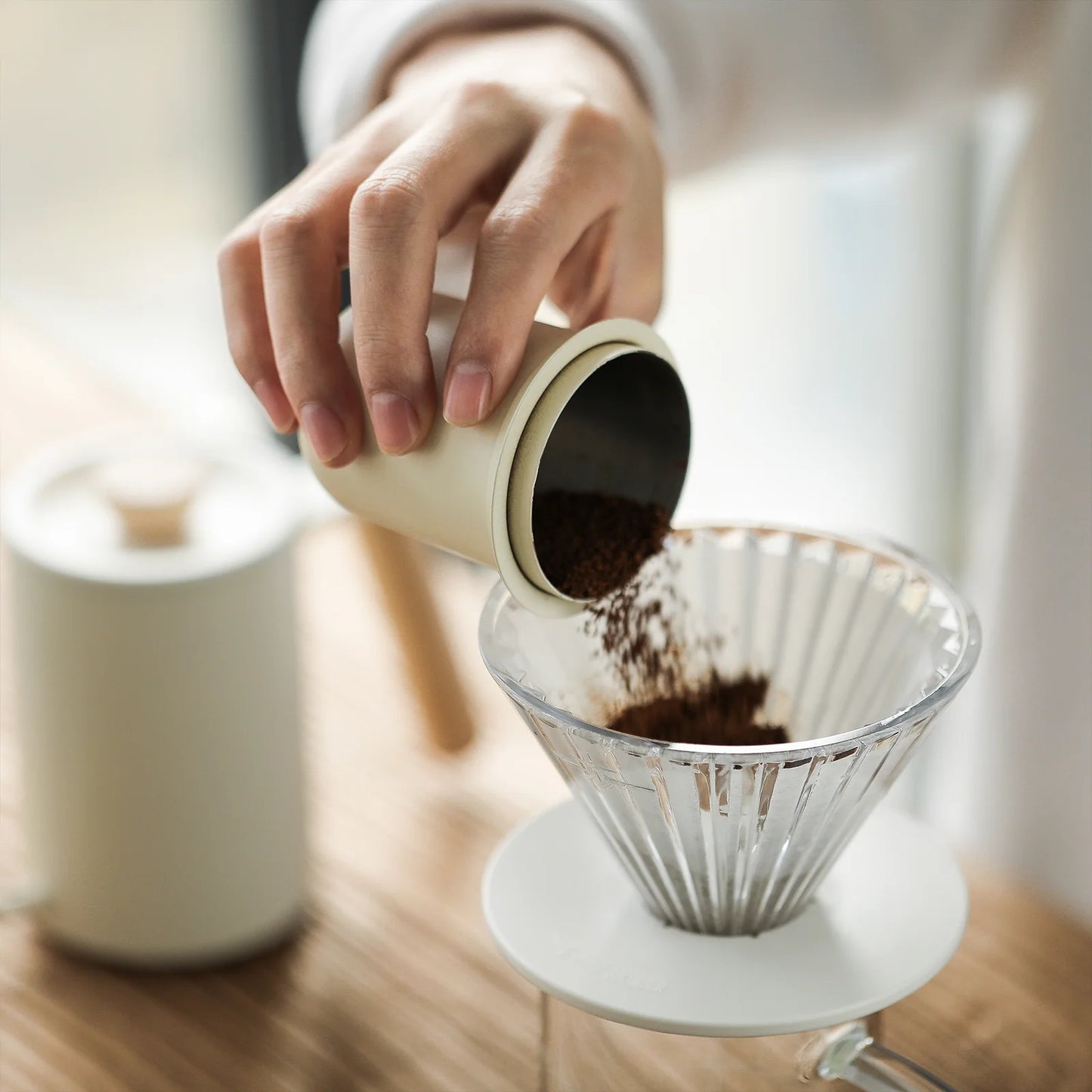 Someone pours ground coffee from a small container into an MHW-3BOMBER Elf Glass Coffee Dripper on a wooden counter. A white canister with a wooden lid sits in the background. The bright, minimalistic scene highlights the drippers elegant triangular diversion groove design.