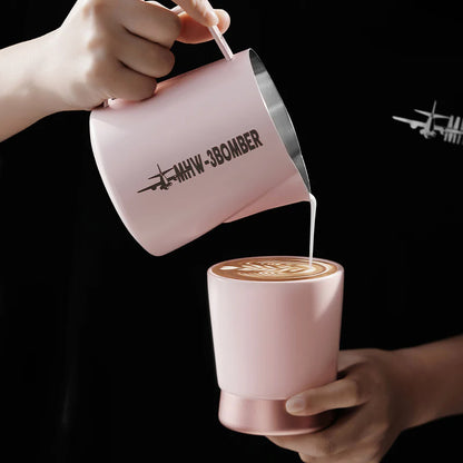 MHW-3BOMBER pink milk pitcher pouring into a pink CERA ceramic cup. Latte art visible. Airplane logo detail on pitcher.
