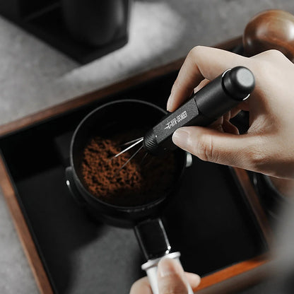 A hand precisely uses the MHW-3BOMBER Lightning Needle Distributor Pro WDT Tool to spread ground coffee in a portafilter on a gray countertop, with coffee accessories and a black tray in the background. Its telescopic design ensures even grounds distribution every time.