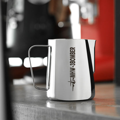 The [Pre-Order] MHW-3BOMBER Latte Art Pitcher 5.0 Glossy, made of 304 stainless steel, has a silver finish with BOMBER in black and features a crocodile spout. It sits on a metallic surface with a blurred red and black background.