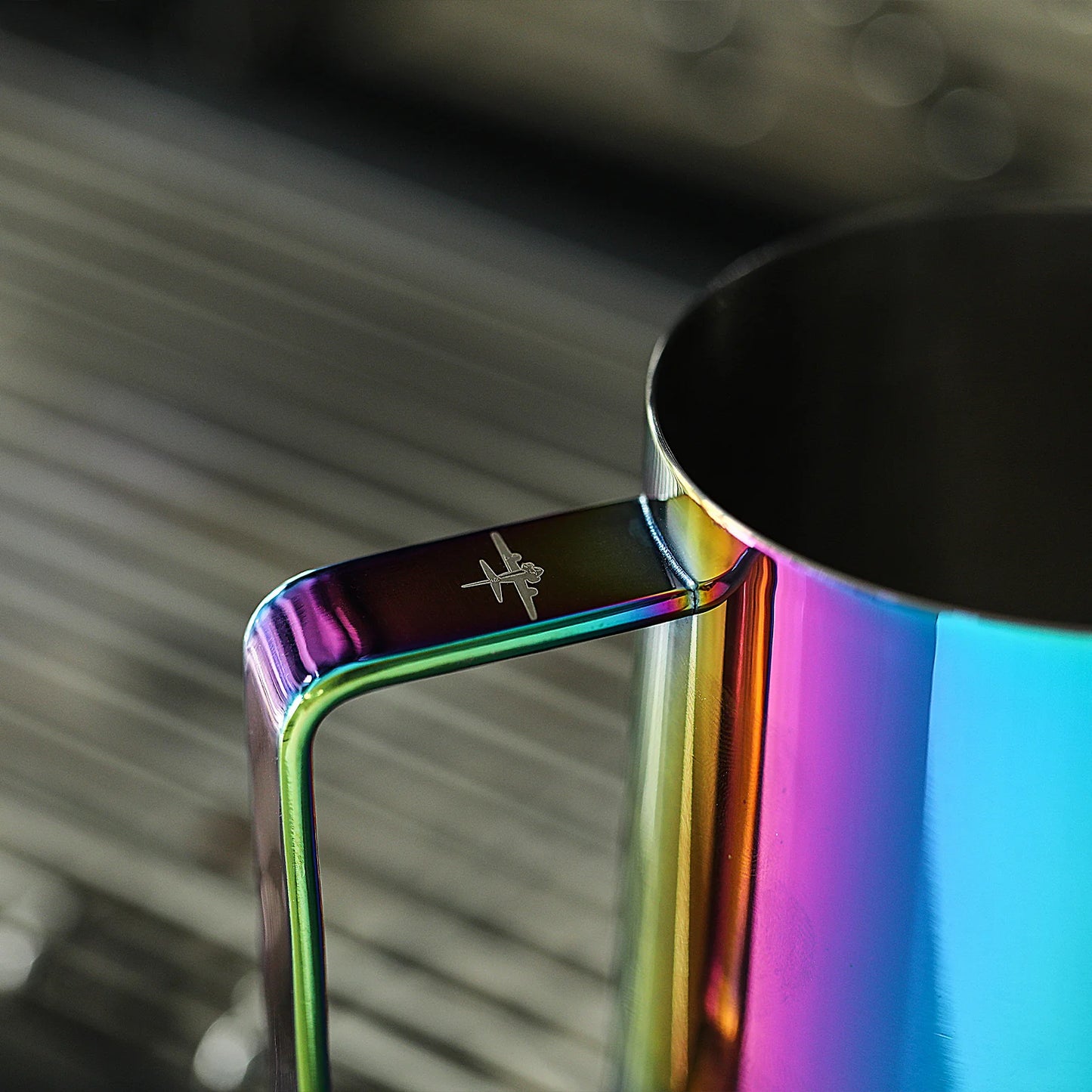 A [Pre-Order] MHW-3BOMBER Latte Art Pitcher 5.0 in an iridescent rainbow finish rests on a silver, ridged surface. The handle has an engraved airplane symbol, and the softly blurred background highlights its allure.